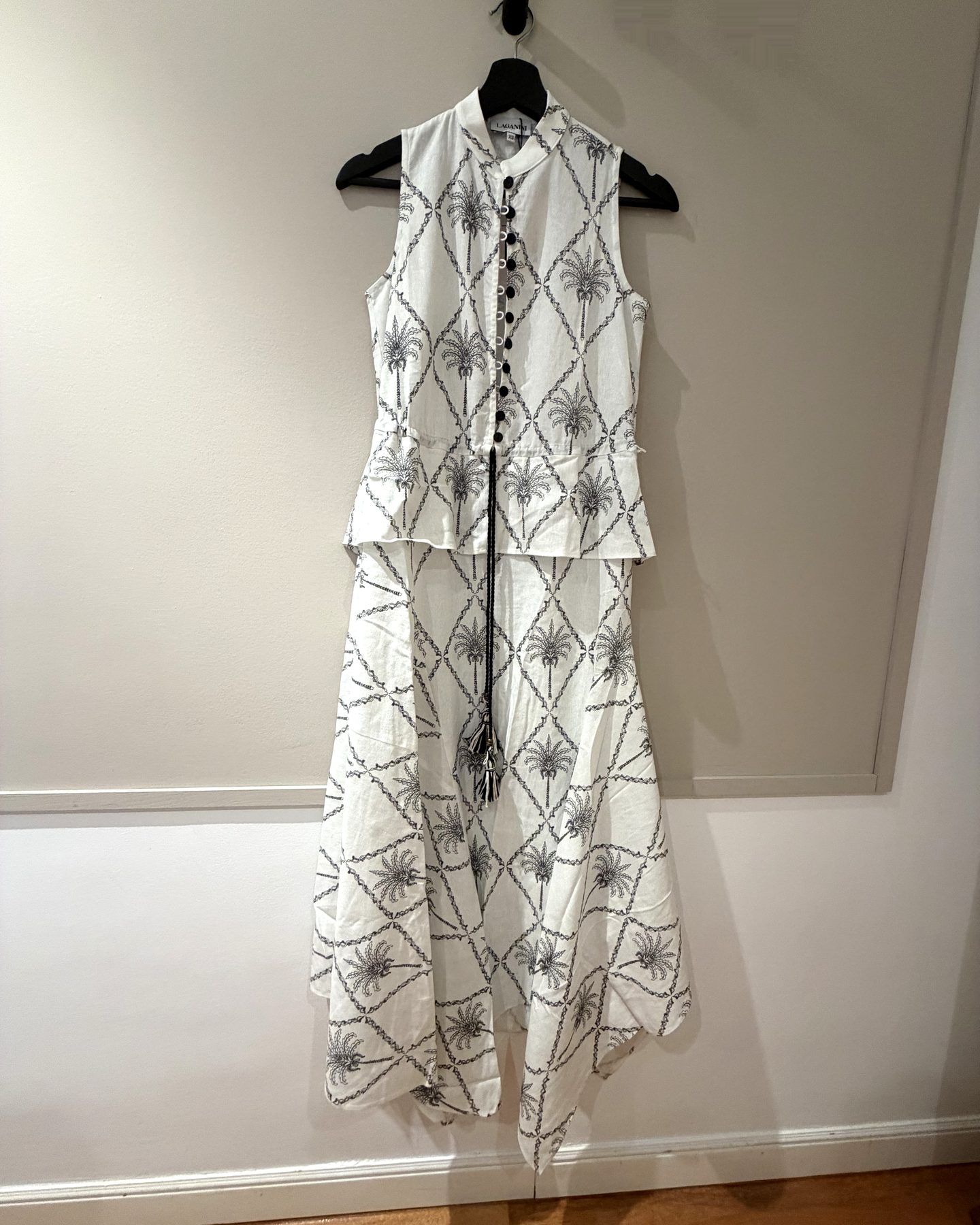 The Ica Hana dress by Laganini Studio — white cotton-linen with a hand-drawn palm-tree print, mandarin collar and buttoned vest bodice — hanging against a cream wall.