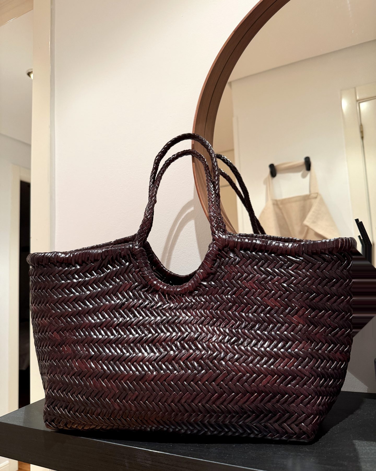 The Matilde bag by Zua — a chocolate-brown hand-braided leather tote with slim braided handles — sitting on a black console in front of a round mirror.